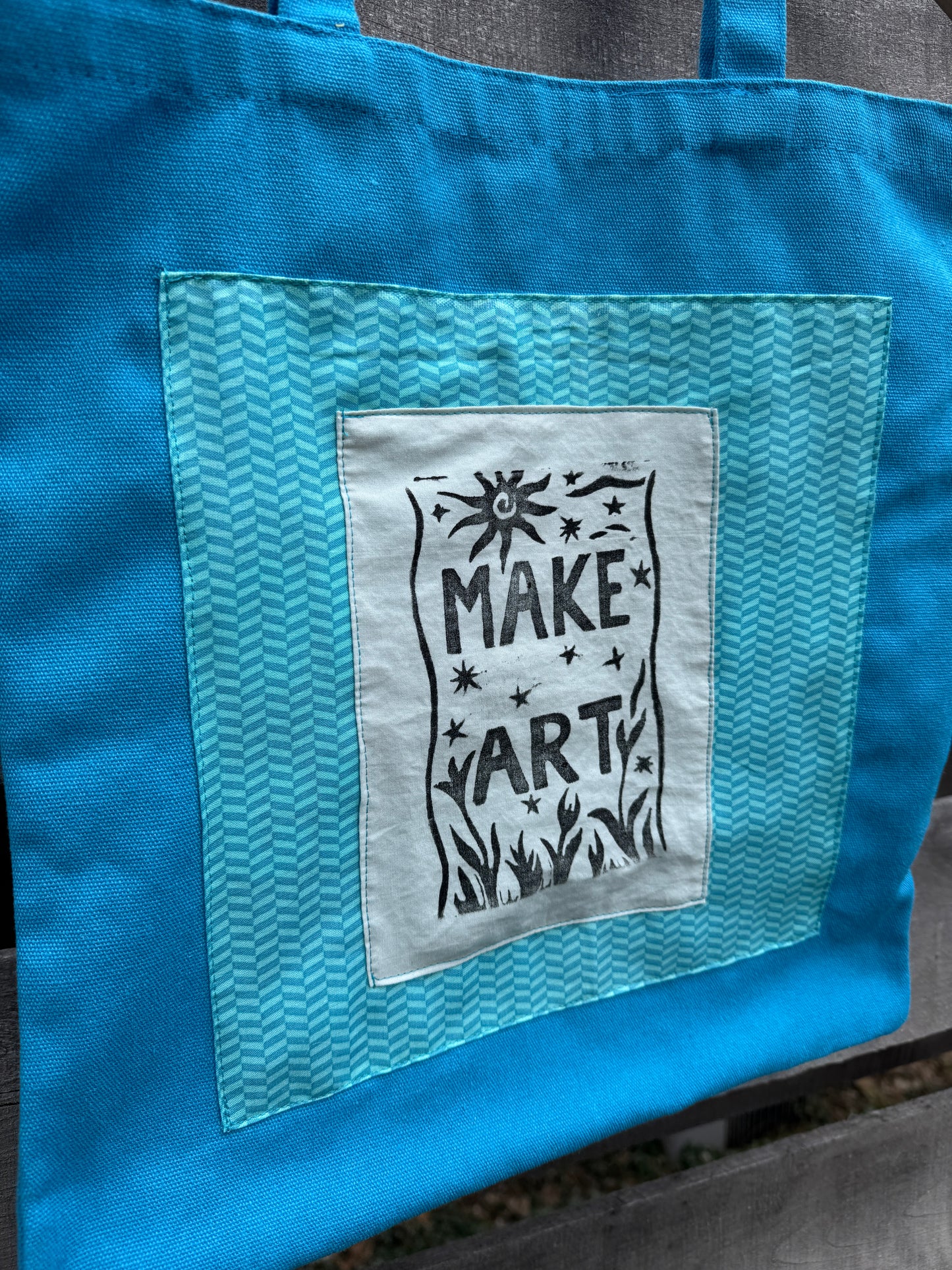 Blue tote bag with a printed "make art" patch on a wooden surface