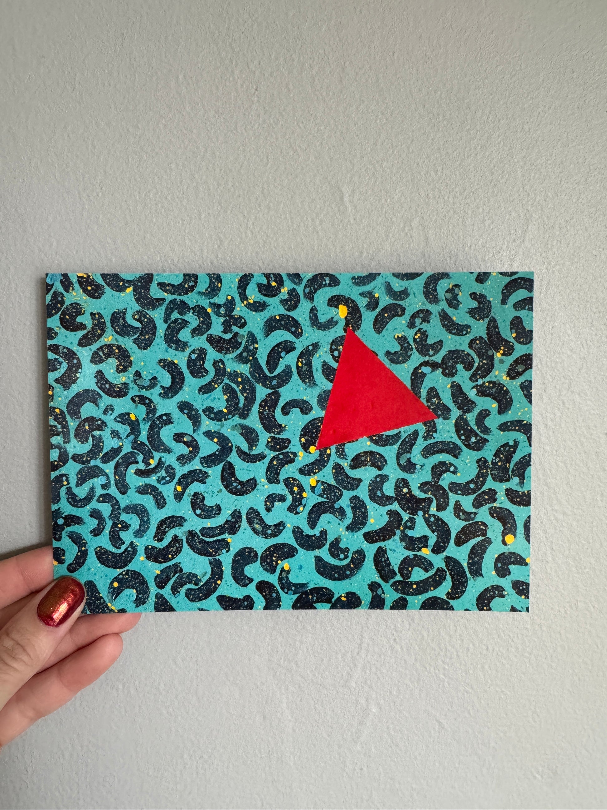 Abstract painting on a postcard held by a hand against a gray wall