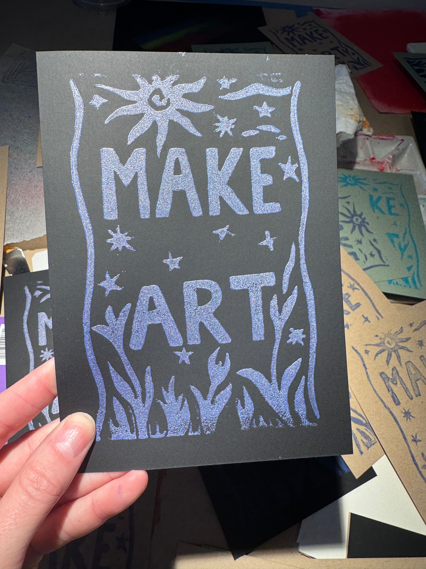 Hand holding a black card with 'MAKE ART' text and decorative elements.