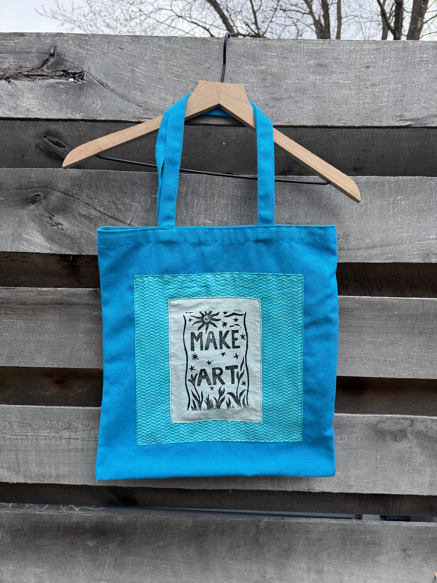 Blue tote bag with a printed "make art" patch on a wooden surface