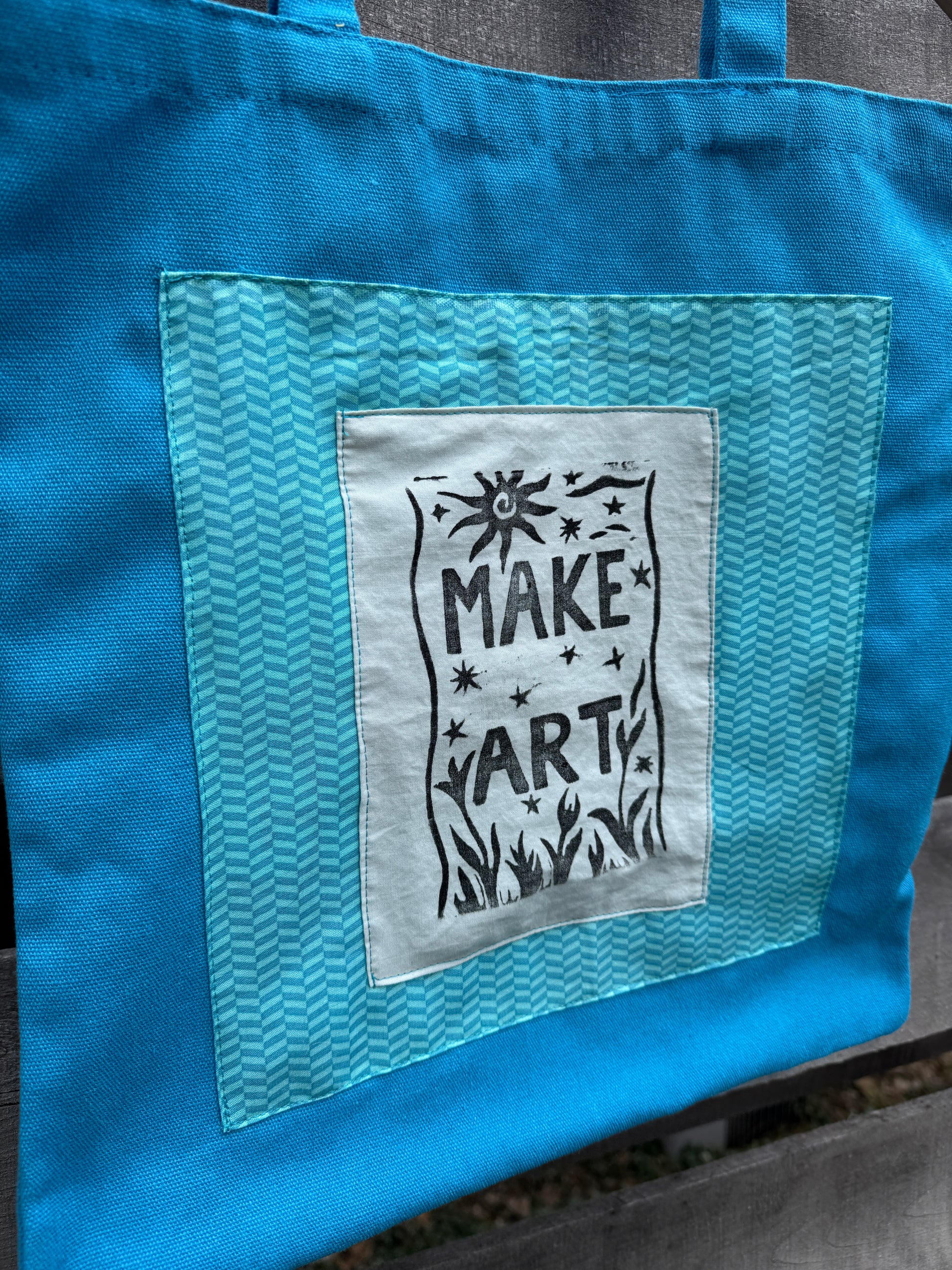 Blue tote bag with a printed "make art" patch on a wooden surface