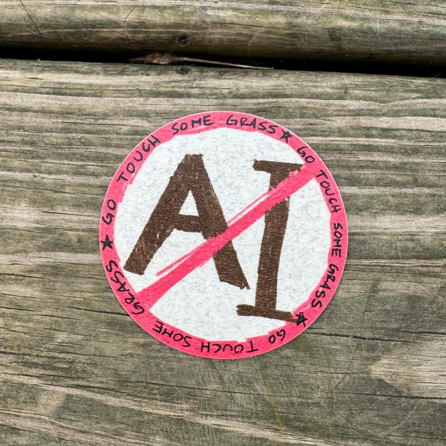 A round sticker with a red and white design featuring the text 'GO TOUCH SOME GRASS' and a red slash over the letters 'AI', placed on a weathered wooden surface.