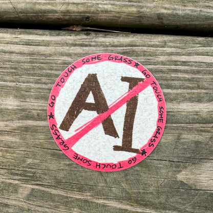 A round sticker with a red and white design featuring the text 'GO TOUCH SOME GRASS' and a red slash over the letters 'AI', placed on a weathered wooden surface.