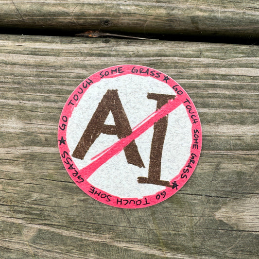 A round sticker with a red and white design featuring the text 'GO TOUCH SOME GRASS' and a red slash over the letters 'AI', placed on a weathered wooden surface.
