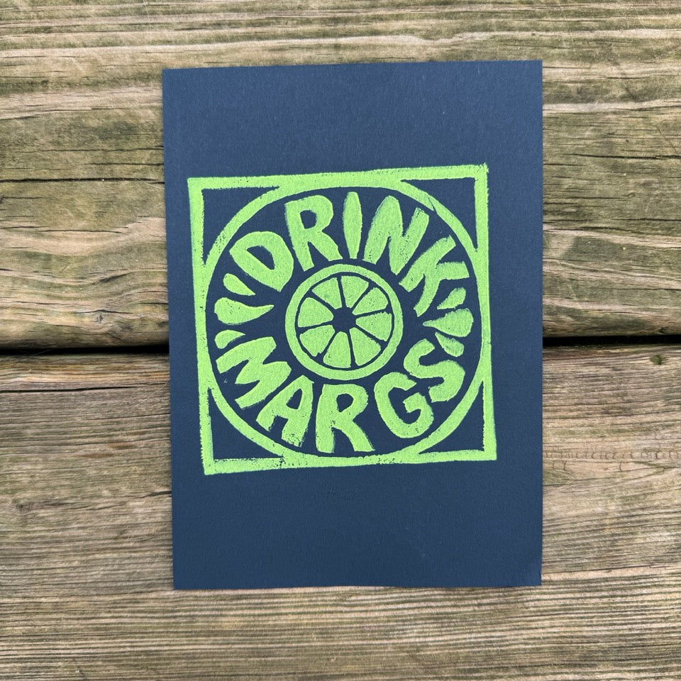 A square navy blue art print or card featuring a lime green block-style logo with the words 'DRINK MARGS' and a circular wedge graphic, photographed flat on a rustic wooden plank surface.
