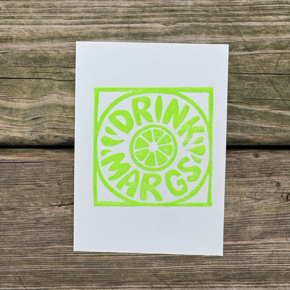 A white rectangular print or card featuring a bright neon green graphic with the words 'DRINK MARGS' and a lime slice motif, placed on a rustic wooden surface.