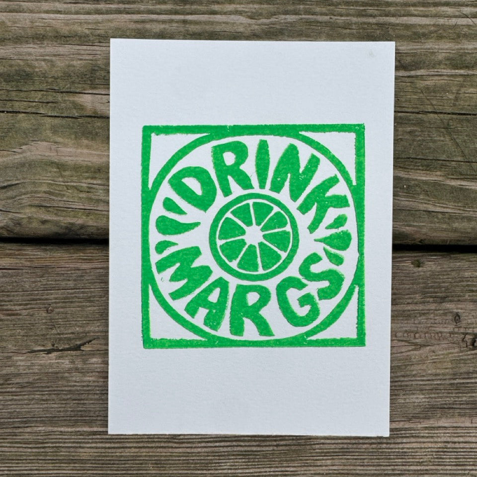 A white art print featuring a green screen-printed parody logo that reads 'DRINK MARGS' in a circular design, displayed flat on a rustic wooden plank surface.