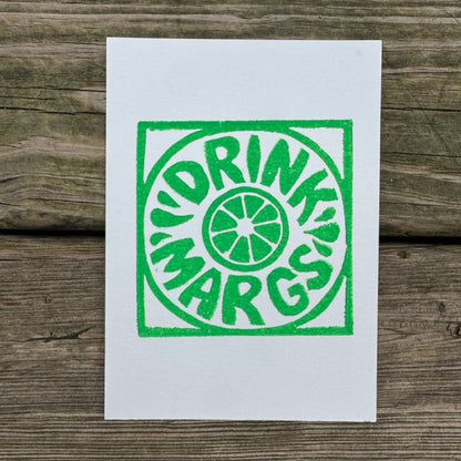 A white art print featuring a green screen-printed parody logo that reads 'DRINK MARGS' in a circular design, displayed flat on a rustic wooden plank surface.