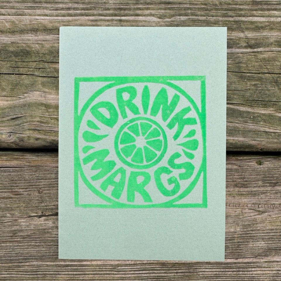A square mint green art print with a green block-print style logo reading 'DRINK MARGS' and a lime slice graphic, displayed flat on a rustic wooden surface.