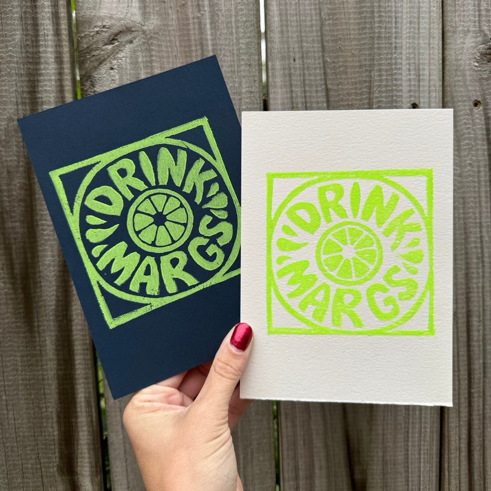 A hand with red nail polish holds two square art prints against a weathered gray wooden fence. One print is dark navy blue with a bright green screenprint, and the other is white with the same bright green screenprint.