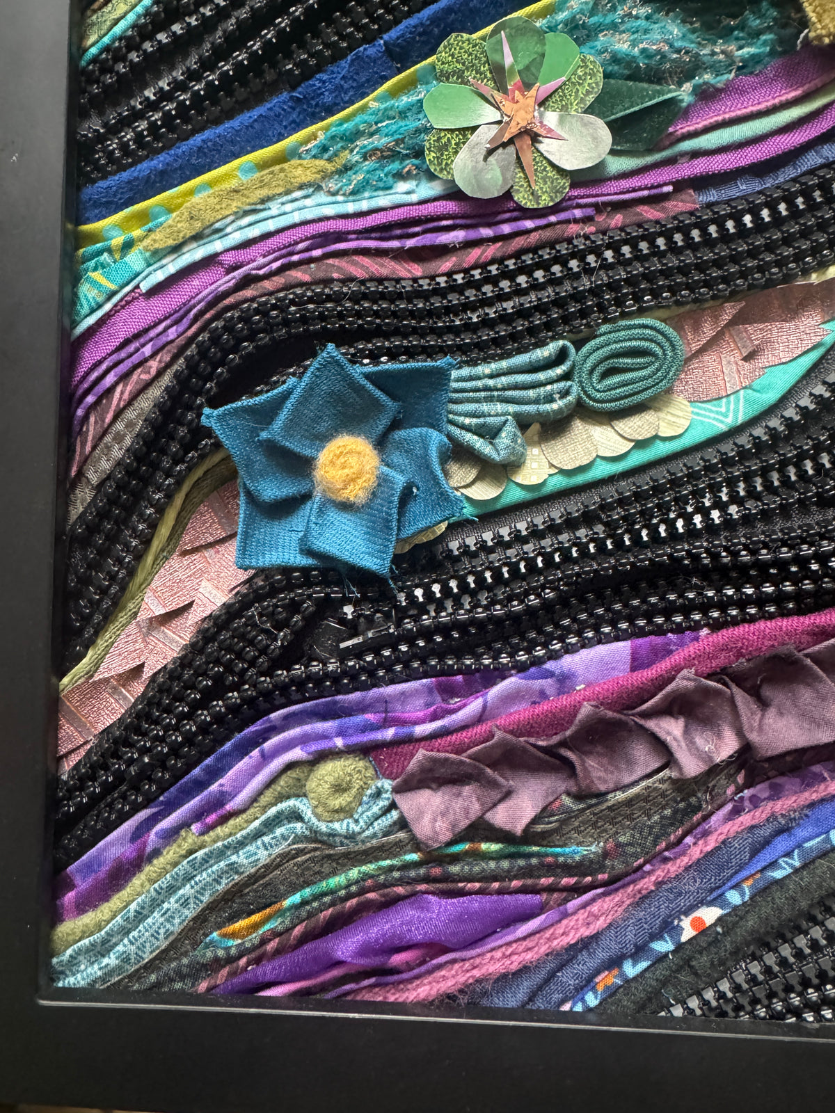 Colorful floral artwork made from fabric strips and zippers in a black frame.